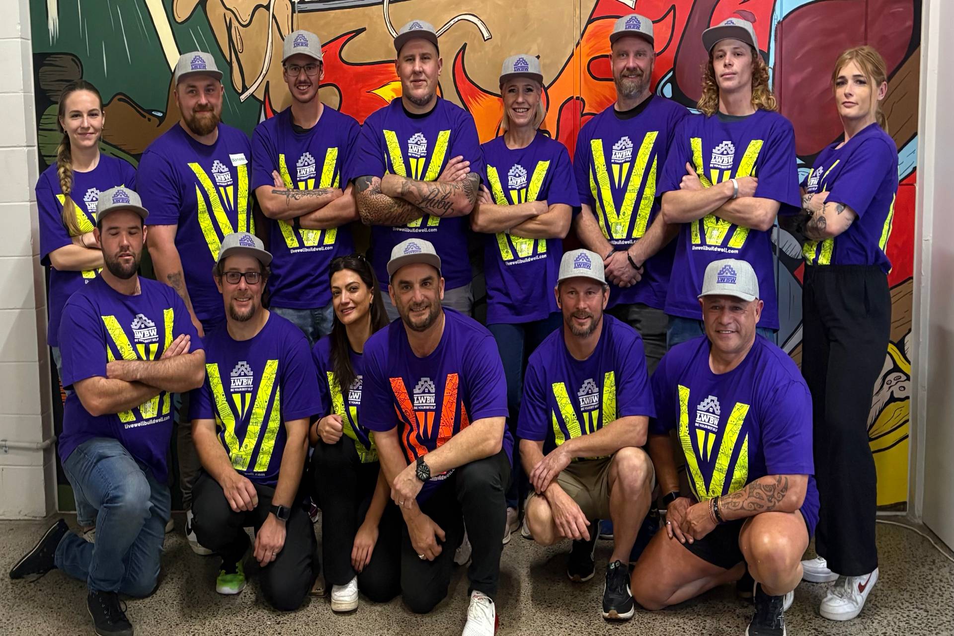A team of people wearing matching purple shirts and gray caps pose together indoors in front of a colorful mural
