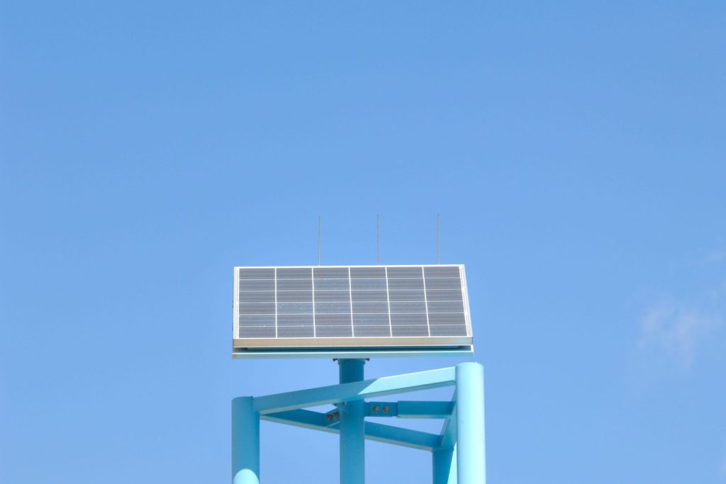 A single, rectangular solar panel with a dark blue surface and metallic grid lines, mounted on a light blue metal frame against a bright, clear sky.